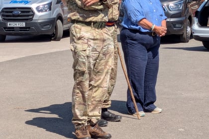 Two people in military uniforms stand next to a person in blue shirt and dark trousers, holding a walking stick, outdoors near parked vans on a sunny day.