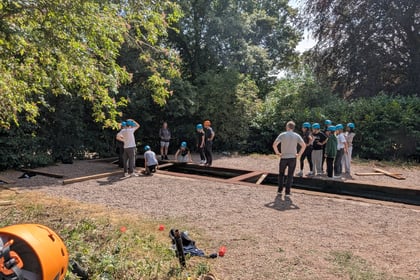 A group of people wearing helmets stand on and around wooden beams outdoors, engaged in a team activity in a sunny, tree-lined area. Equipment and bags are scattered on the ground nearby.
