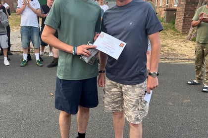 A cadet in civilian clothing holds a certificate which has been presented to him by the adult standing next to him. Other cadets are in the background applauding.