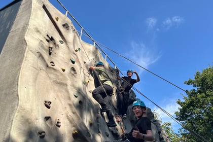 One cadet punches the air at the top of a climbing wall, where another is still finishing the climb. A third cadet nearer the bottom of the wall looks at the camera and also has a fist clenched.