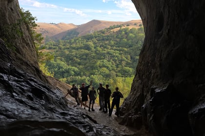 A view from inside a cave looking out into the vista of trees and hills. Six cadets are silhouetted in the entrance to the cave.