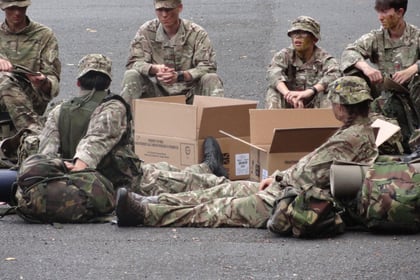 Cadets relaxing in their camouflage uniform around two large cardboard boxes which are on the ground in the middle of them