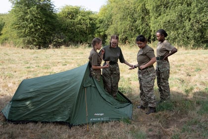 Four cadets in uniform are chatting outside the entrance to a green tent. They are standing in a field with trees in the background.