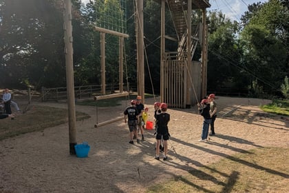 Four children in helmets stand by a tall outdoor climbing structure with wooden poles, ropes, and a platform. Two adults supervise. There are trees and grass around, under a sunny sky.