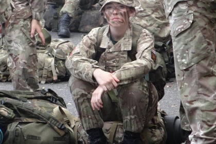 A cadet looks at the camera in full uniform, siiting on their backpack. Other cadets are in the background doing the same.