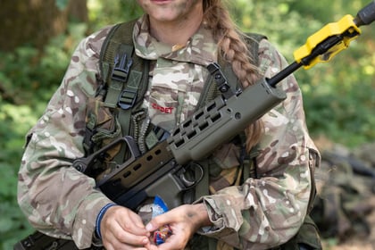 A cadet in camouflage smiles at the camera, holding a weapon across her front and a bag of haribo in her hands
