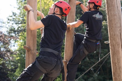 two cadets on a wooden climbing apparatus in safety gear, from a behind view