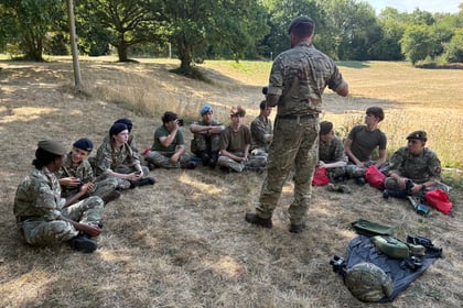 Cadets in camouflage sit in a semi circle around an instructor with his back to the camera. Some kit is laid on the ground for demonstration