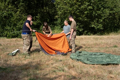 Two cadets at either end of a tent canvas and one in the middle, putting the tent pole into the canvas to build it.