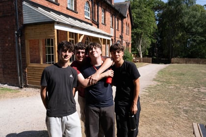 Four young people pose for the camera outside a brick building. One is standing slightly behind with his arms around another.