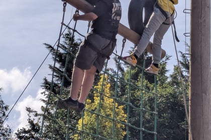 Two people wearing helmets and harnesses climb an outdoor adventure course with a rope net and tyres, surrounded by trees under a partly cloudy sky.