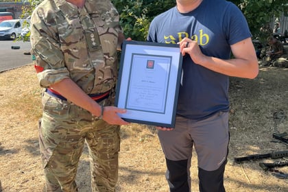 Two men stand outside on dry grass. One wears military uniform and glasses, the other wears a casual t-shirt and trousers. They smile and hold a framed certificate together whilst posing for the photo. Green bushes are visible in the background.