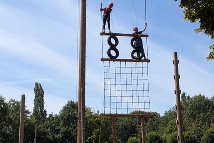 A group of people in helmets watch as others climb a tall outdoor ropes course with nets, tyres, and wooden beams, surrounded by trees and blue sky.