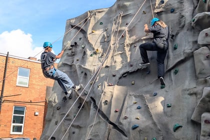 Several people wearing blue helmets and harnesses are climbing an outdoor rock wall, whilst others watch from below. The scene is set in a sunny area with buildings in the background.