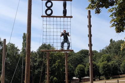 A group of people in helmets stand near a tall outdoor climbing structure. One person is climbing a rope net, whilst others watch from below. Trees and a fence are visible in the background.