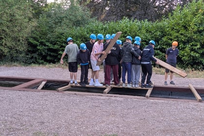 A group of children in blue helmets balance on wooden planks over a shallow trench outdoors, working together as a team-building activity. One child in an orange helmet stands apart, holding a plank. Green bushes are in the background.