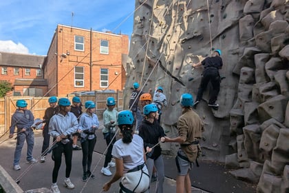 A group of people wearing helmets stand by a climbing wall outside, as one person climbs and others watch or assist. Brick buildings and a blue sky are in the background.