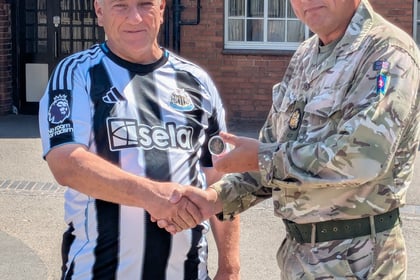 A man in a Newcastle United football shirt shakes hands with a man in military uniform outside a brick building. The man in uniform hands over a small round object.