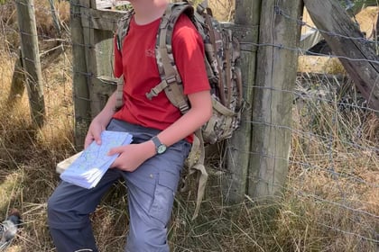 A boy wearing sunglasses, a red shirt, and a rucksack sits on a wooden fence holding a map, surrounded by dry grass and countryside scenery.
