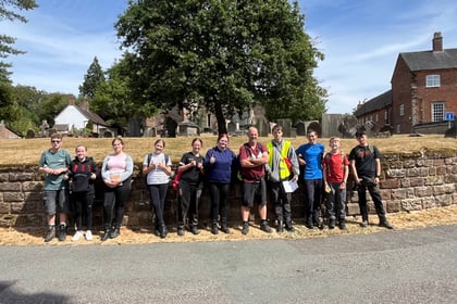 A group of thirteen people stand in a queue outside on a sunny day, posing in front of a stone wall, trees, and brick buildings. Most are wearing casual or hiking clothing and smiling at the camera.