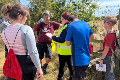 A group of people with rucksacks stands outdoors on dry grass, looking at papers or maps. One person wears a hi-vis vest, and the sky is blue with some trees in the background.