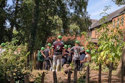 A group of people wearing green helmets gather outdoors in a wooded area near a building on a sunny day.