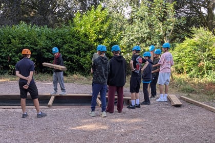 A group of young people stand by a pit with logs of wood to use to cross it.
