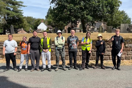Nine young people stand side by side in front of a stone wall and trees, some wearing high-visibility vests. Most are smiling, dressed casually, and the weather appears sunny and clear.