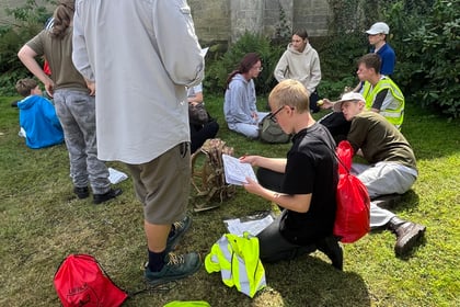 A group of people, some in hi-vis vests, sit and stand on grass outside an old stone building, reading papers and talking. Red rucksacks and jackets are on the ground near them.