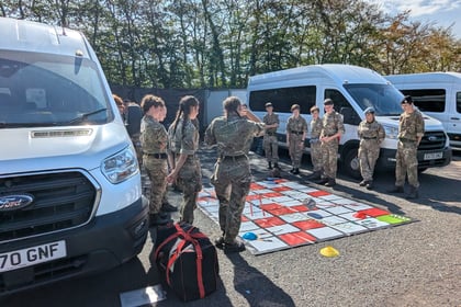A group of people in military uniforms stand around a large board game mat on the ground, between several white vans, in a sunny outdoor setting.