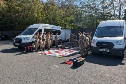 A group of people in military uniforms gather around a large mat with a grid pattern, outdoors between two white vans, with trees and parked vehicles in the background.