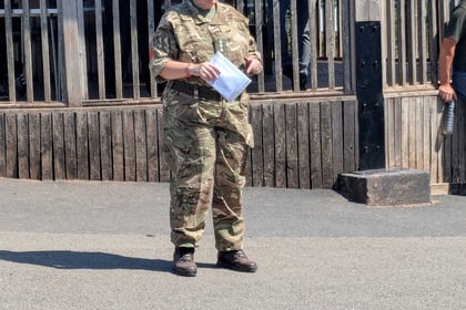 A person in military uniform stands on pavement holding papers, speaking to others. Three people stand behind a wooden railing under a shaded structure, watching the speaker.