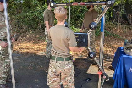 A boy in camouflage trousers and a brown shirt uses a light reaction game outdoors, under a canopy, with other people in similar attire nearby. A blue table with a helmet is to the right.