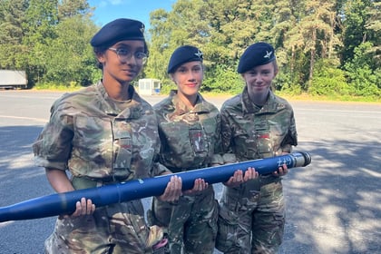 Three young women in camouflage uniforms and berets stand outdoors, holding a large blue cylindrical object together, with trees in the background.