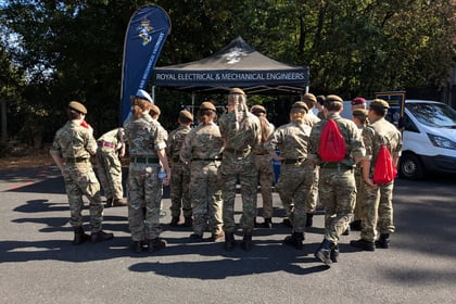 A group of people in camouflage uniforms stand facing a tent labelled Royal Electrical & Mechanical Engineers, with trees and a white van in the background.