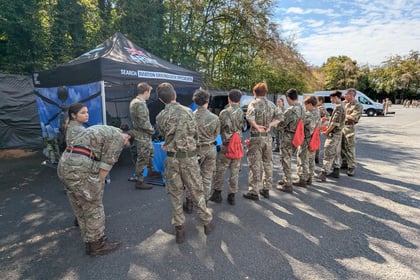 A group of people in camouflage uniforms stand in a queue near a tent labelled SEARCH BAVARIAN GROUNDHOG SPECIALIST, with trees and a parked van in the background.