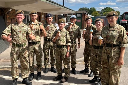 Eight people in military uniforms stand outdoors on a sunny day, smiling and holding ice cream cones. Trees, a building, and a partly cloudy sky are visible in the background.