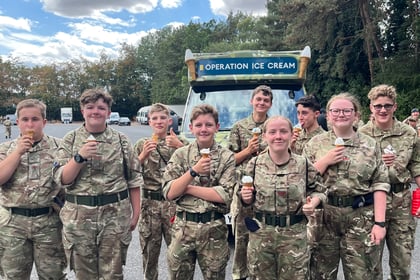 A group of people in camouflage uniforms stand outdoors, smiling and holding ice cream cones in front of a sign that says Operation Ice Cream. Trees and cars are visible in the background.