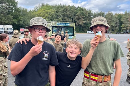 Three smiling teenagers in military-style uniforms enjoy ice cream outdoors, with one boy standing in the middle and hugging the other two. Other people in similar uniforms and trees are in the background.