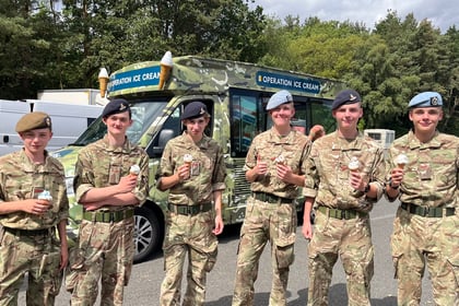 Six people in military uniforms stand in front of a camouflage ice cream van, each holding an ice cream cone, smiling outdoors on a sunny day with trees in the background.