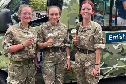 Three young people in camouflage uniforms stand smiling in front of an ice cream van, each holding an ice cream cornet. The van has a camouflage pattern and a sign that reads British.