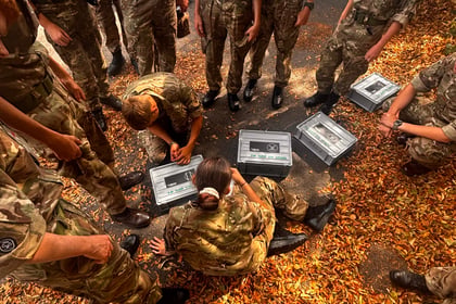 A group of young people in camouflage uniforms sit and stand on leaf-covered ground, opening metal cases. Some are focused on the cases, whilst others look on.