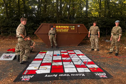 Five people in military uniforms stand around a large Know Your Army game mat on the ground near a brown skip and trees. Dice, ladders, and other game elements are visible on the mat.