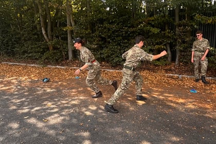 Two people in camouflage uniforms run between blue cones on a paved area surrounded by trees, whilst a third person stands nearby watching.