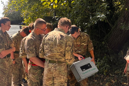 A group of people in camouflage uniforms stand together outdoors, looking at a grey box. They are near trees and sunlight filters through the leaves.