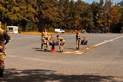 Four people in military uniforms place orange traffic cones on an open paved area with trees and parked vehicles in the background under a blue sky.