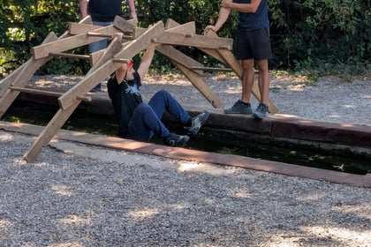 Three people participate in an outdoor activity. Two stand on a wooden bridge, one wearing an orange helmet. The third person hangs underneath the bridge over a narrow water channel. Trees and gravel surround them.