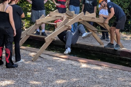 A group of people in helmets stand around a wooden structure over a small stream while one person crawls underneath it. The scene appears to be part of an outdoor team-building activity.