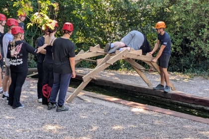 A group of people wearing helmets assist someone crawling over a wooden bridge structure outdoors, with trees and a narrow water channel nearby.