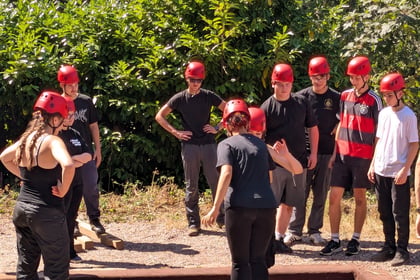 A group of people wearing red helmets gather outdoors, watching one person cross a narrow wooden beam over a shallow pit. Trees and bushes are in the background.